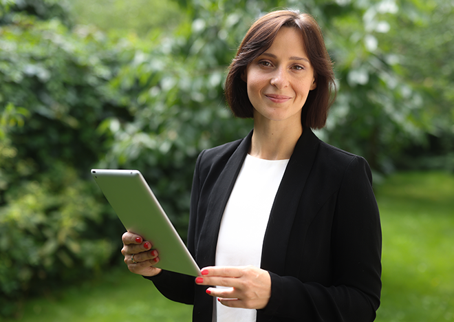 Kobieta - audotorka ESG, w formalnym stroju trzymająca tablet, stojąca na tle zieleni.