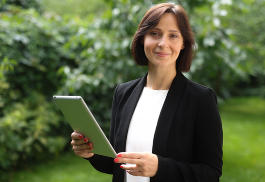 Kobieta - audotorka ESG, w formalnym stroju trzymająca tablet, stojąca na tle zieleni.